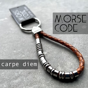 May include: Brown leather keychain with a silver metal ring and a silver metal bead chain. The keychain has the text "MORSE CODE" on it and the text "carpe diem" on the bottom.