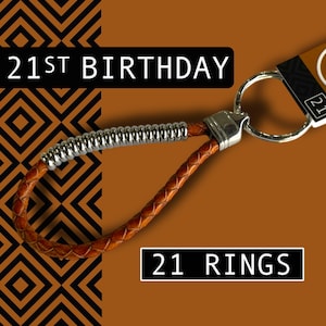 May include: A brown leather keychain with a silver metal accent and a silver key ring. The keychain has the text "21ST BIRTHDAY" and "21 RINGS" on a brown background with a black and orange geometric pattern.