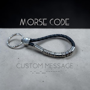 May include: A black braided leather keyring with silver-coloured metal beads and a key ring. The words "MORSE CODE" and "CUSTOM MESSAGE" are above and below the keyring. The Morse code for the custom message is also displayed.