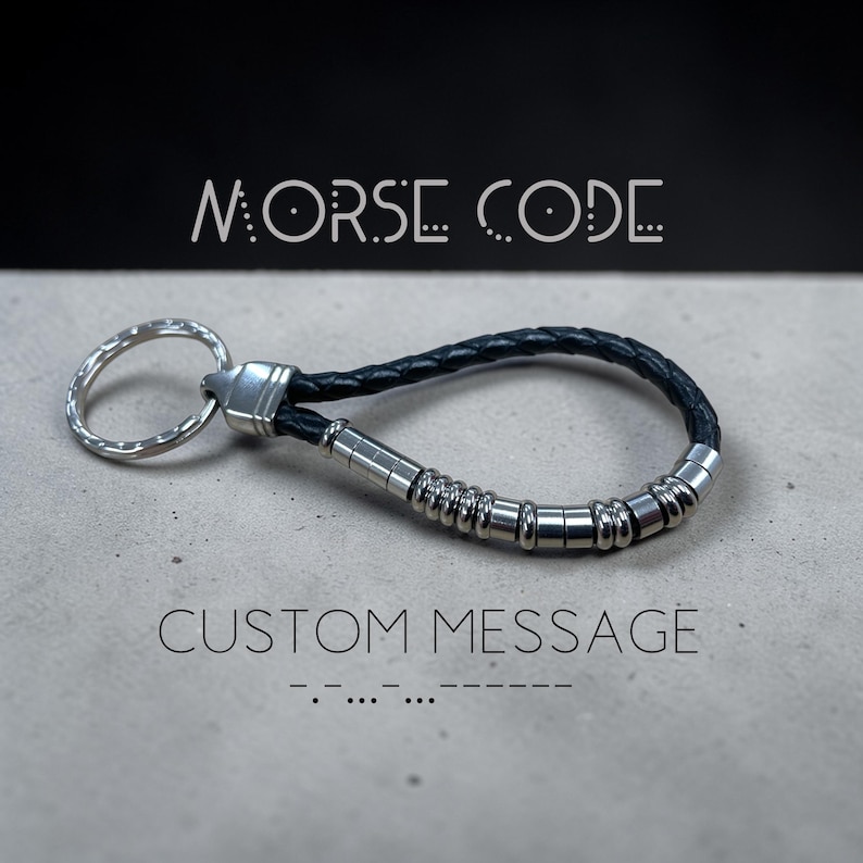 K&ouml;nnte beinhalten: Ein Schl&uuml;sselanh&auml;nger aus schwarzem geflochtenem Leder mit einem silberfarbenen Metallring und einer Reihe kleiner, zylindrischer silberfarbener Perlen entlang der Lederschlaufe. Der Text MORSE CODE und CUSTOM MESSAGE ist sichtbar.