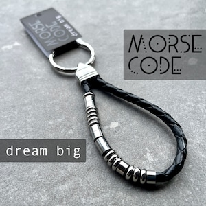 May include: Black leather keychain with a silver ring and a silver chain of beads. The keychain has the words "MORSE CODE" and "dream big" printed on it.