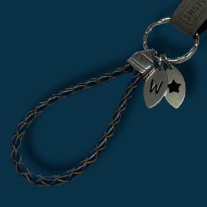 May include: A blue braided leather keychain with silver-tone metalwork. The keychain has two silver charms, one with the letter "W" and the other with a star cutout. It's attached to a dark grey leather strap and a silver keyring.