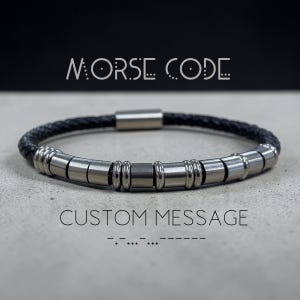 May include: A black braided leather bracelet with silver cylindrical beads. The bracelet features a magnetic clasp. The words "MORSE CODE" and "CUSTOM MESSAGE" are printed above and below the bracelet.