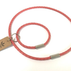 May include: Two braided leather cords in a pink color. The larger cord is a necklace with a silver clasp. The smaller cord is a bracelet with a silver clasp. The cords are on a white background.