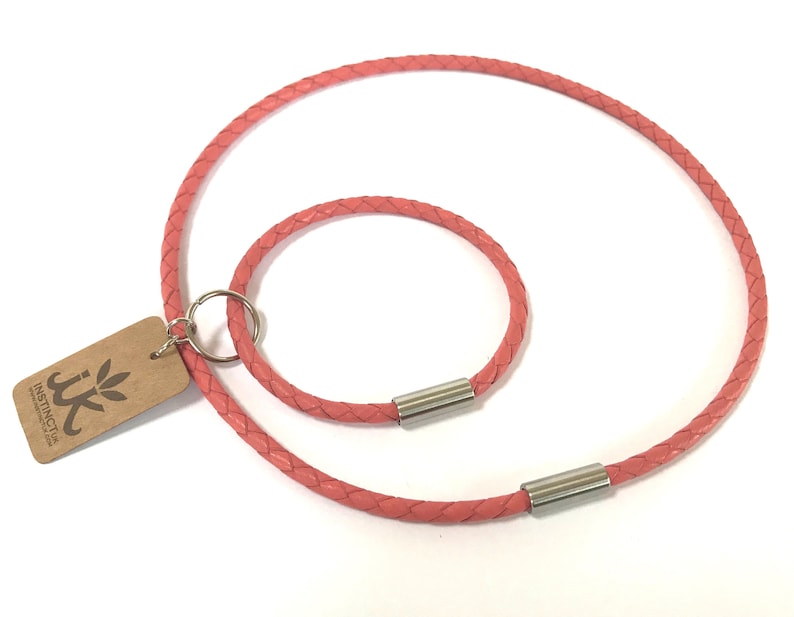 May include: Two braided leather cords in a pink color. The larger cord is a necklace with a silver clasp. The smaller cord is a bracelet with a silver clasp. The cords are on a white background.