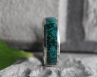 May include: A silver ring with a wide band featuring a vibrant turquoise stone inlay. The stone has a mottled appearance with shades of green and dark blue. The ring is set against a textured gray background.