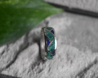 May include: A silver ring with a geometric pattern of teal and blue inlays. The ring is set against a light gray, textured surface, with a green leaf in the background. The ring's design features a repeating pattern.