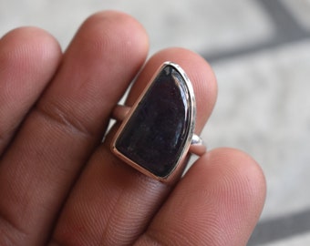 Può includere: Un anello d'argento con una pietra preziosa di colore viola scuro. La pietra preziosa è incastonata in una fascia d'argento e ha una forma leggermente curva.