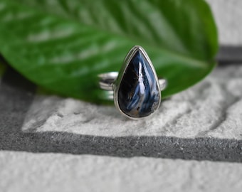 Op de afbeelding: Een zilveren ring met een druppelvormige edelsteen. De steen heeft een donkerblauw en wit patroon, gezet in een gepolijste zilveren band. De ring wordt gepresenteerd op een getextureerd grijs oppervlak met een groen blad op de achtergrond.