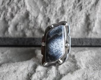May include: A silver ring featuring a large, oval-shaped, black and white dendritic agate stone set in a silver bezel. The ring has a textured, organic design.