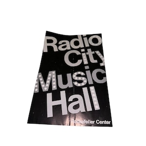 May include: A black and white Radio City Music Hall promotional flyer. The words "Radio City Music Hall" are prominently displayed in large white font. The words "Rockefeller Center" are at the bottom.