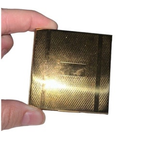 May include: A gold-colored metal compact with a textured surface and a raised rectangular area in the center.