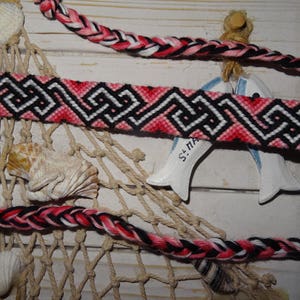 Puede incluir: Un brazalete de amistad tejido en rosa, negro y blanco con un patrón geométrico. El brazalete está sobre una superficie blanca con un material de red y conchas.