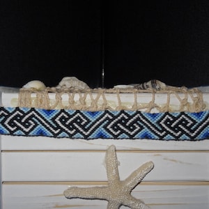 May include: A blue, black, and white woven friendship bracelet with a geometric pattern. The bracelet is displayed on a white wooden surface with a starfish.