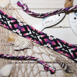 Peut inclure: Un bracelet d'amitié noir, rose, blanc et violet avec un motif géométrique. Le bracelet est posé sur une surface en bois blanc avec un filet de pêche et deux poissons en céramique blanche.