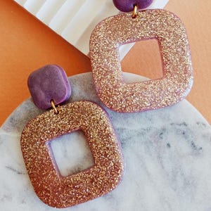 May include: A pair of purple and gold glitter square earrings with a square cutout in the center. The earrings have a purple top and a gold glitter bottom.