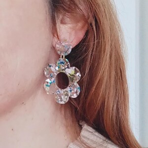 May include: A pair of clear flower-shaped earrings with a gold-tone metal post and a smaller clear flower charm hanging from the bottom. The earrings are decorated with iridescent glitter.