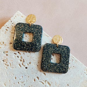 Op de afbeelding: Een paar vierkante oorbellen met een gouden accent. De oorbellen zijn zwart met gouden glitter.