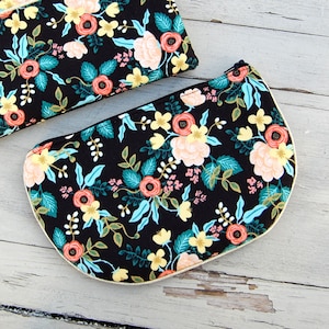 Puede incluir: Dos bolsas de tela negras con un estampado floral en tonos rosa, amarillo y verde. Las bolsas tienen cierre de cremallera y un ribete de color crema.