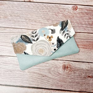 Op de afbeelding: Een lichtblauwe en witte bloemenclutch met een klep sluiting. De clutch is gemaakt van stof en heeft een bloemenpatroon met blauwe, witte en bruine bloemen. De clutch ligt op een houten oppervlak.