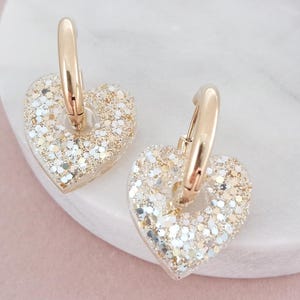 Könnte beinhalten: Ein Paar goldfarbene Creolen mit herzförmigen Anhängern. Die Herzanhänger sind mit glitzernden Gold- und Silberpartikeln gefüllt. Die Ohrringe liegen auf einer weißen Marmoroberfläche vor einem zartrosa Hintergrund.