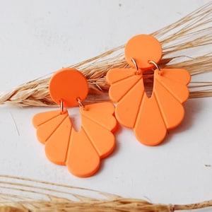 Könnte beinhalten: Auffällige orangefarbene Ohrringe. Jeder Ohrring hat einen kreisförmigen oberen Teil und einen fächerförmigen unteren Teil mit vertikalen Linien. Die Ohrringe bestehen aus einem glatten Material und werden von goldfarbenen Haken gehalten. Der Hintergrund umfasst Weizenähren.