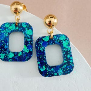 Puede incluir: Un par de pendientes de forma cuadrada con una parte superior de metal dorado y un diseño de purpurina azul y verde.