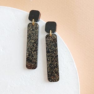 Peut inclure: Une paire de boucles d'oreilles pendantes noires et dorées à paillettes avec un dessus carré et un bas rectangulaire.