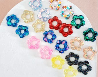 Könnte beinhalten: Sortiment von 18 bunten Glitzer-Ohrringen in Blumenform aus Harz. Jeder Ohrring hat ein kleines Loch in der Mitte. Die Ohrringe sind in einem Halbkreis auf einer weißen Oberfläche angeordnet.