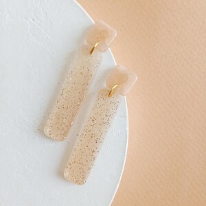 Op de afbeelding: Een paar lange, rechthoekige hangende oorbellen met een lichtbruine, doorschijnende basis en gouden glitter. De oorbellen hebben een klein vierkant stukje bovenaan.