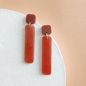 Puede incluir: Un par de pendientes colgantes con una parte superior cuadrada marrón y una parte colgante rectangular marrón larga. Los pendientes tienen una textura brillante y brillante.