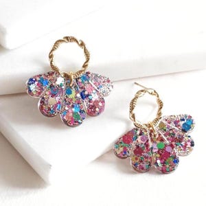 May include: A pair of dangle earrings with a floral design. The earrings feature a gold-colored twisted hoop and a flower-shaped pendant filled with colorful glitter. The glitter includes shades of pink, blue, green, and purple.
