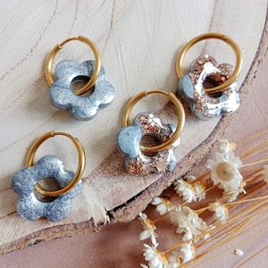 Op de afbeelding: Vier paar bloemvormige oorbellen met goudkleurige hoepels. Twee paar hebben een gemarmerde grijze uitstraling, terwijl de andere twee een grijze basis hebben met gouden glitteraccenten. De oorbellen worden gepresenteerd op een houten oppervlak.