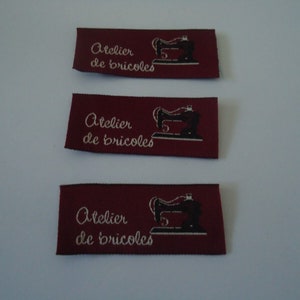 Peut inclure: Trois étiquettes en tissu bordeaux avec du texte blanc qui dit "Atelier de bricoles" et une illustration en noir et blanc d'une machine à coudre.
