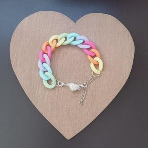 Peut inclure: Un bracelet chaîne coloré avec des maillons pastel roses, oranges, jaunes, verts et bleus. Le bracelet est doté d'un fermoir argenté avec un charm en forme de cœur et d'une chaîne réglable. Il est présenté sur une surface en forme de cœur en bois.