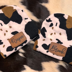 May include: Two cow print knit beanies with brown leather patches. The patches have the names "Reagan" and "Brook" written in black.