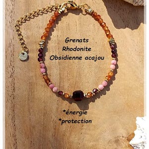 Peut inclure: Un bracelet composé de perles facettées orange, rouge foncé et rose, avec une perle carrée centrale rouge foncé, des intercalaires dorés et une chaînette d'extension dorée. Le texte indique : Grenats, Rhodonite, Obsidienne acajou, *énergie, *protection.
