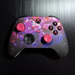 May include: A black and pink Xbox controller with a galaxy-like design. The controller has pink buttons and a pink d-pad.