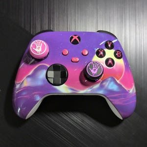 May include: A wireless video game controller featuring a vibrant purple and pink nebula-inspired design with mountain silhouettes. It includes black thumbsticks with pink handprint icons, a black D-pad, and buttons labeled X, Y, A, and B.