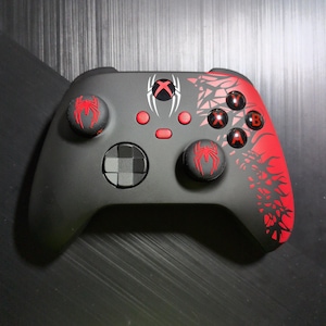 May include: A matte black wireless video game controller featuring red spider icons on the analog sticks, a red and white spider emblem above the center buttons, and a red geometric web pattern on the right side with red A, B, X, and Y buttons.
