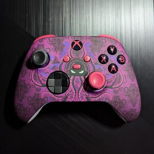 May include: A pink and purple Xbox controller with a black octopus design. The controller has pink buttons and a black faceplate.