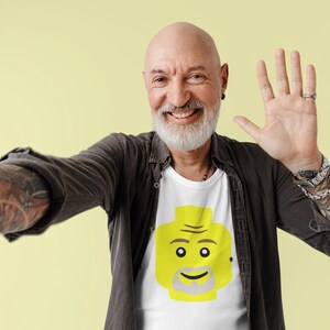 May include: A man wearing a black shirt with a white t-shirt underneath. The t-shirt has a yellow Lego-style face on it. He is smiling and waving with his right hand.