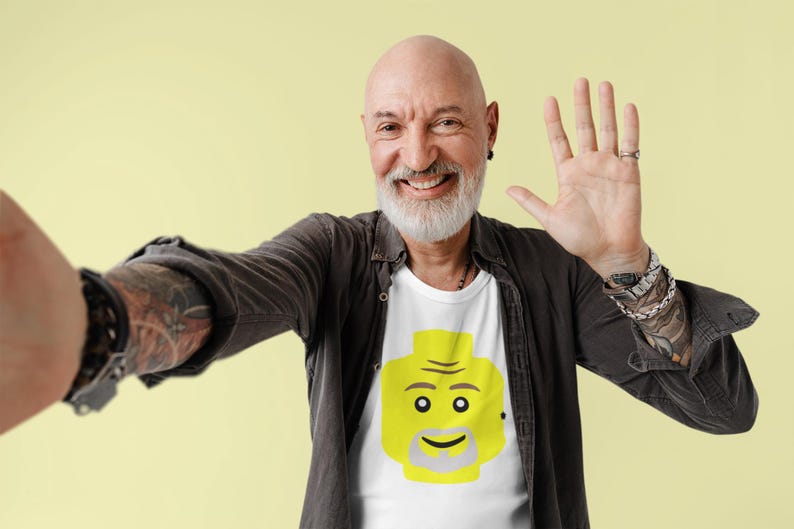 May include: A man wearing a black shirt with a white t-shirt underneath. The t-shirt has a yellow Lego-style face on it. He is smiling and waving with his right hand.