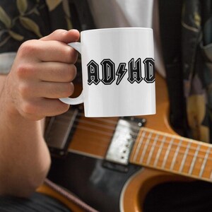 Man holding white mug printed with black AD HD logo while playing electric guitar, showcasing ADHD SVG cut file for mugs or gifts.