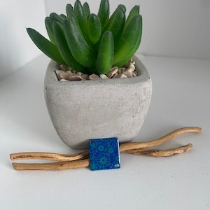 Peut inclure: Une petite plante succulente verte dans un pot en béton gris rempli de petits galets. Un carreau décoratif bleu carré avec un motif floral repose sur deux morceaux de bois flotté.