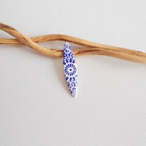 Może przedstawiać: Biały ceramiczny wisiorek z niebiesko-białym geometrycznym wzorem. Wisiorek ma kształt łzy i ma otwór u góry do zawieszenia.