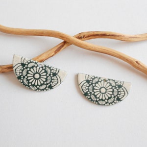 Może przedstawiać: Dwa ceramiczne kolczyki w kształcie półkola z kwiatowym wzorem w kolorze ciemnozielonym i białym. Kolczyki umieszczone są na białej powierzchni z zakrzywioną drewnianą gałęzią. Kolczyki mają otwór do zawieszenia.