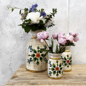 May include: Three cream-coloured vases with floral designs and yellow rims hold fresh flowers. The largest vase contains white and blue flowers, while the smaller vases display pink peonies. The vases sit on a wooden table.
