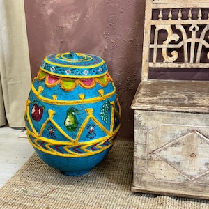 May include: A large, blue, egg-shaped ceramic pot with a lid. The pot is decorated with yellow, red, green, and white painted designs of fruits and flowers. The pot is sitting on a brown woven rug.