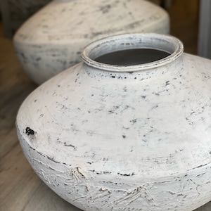 May include: Two large, white, distressed ceramic vases with a rounded shape and wide openings. The vases have a textured, aged appearance with dark accents, suggesting a rustic or vintage style. The background is blurred, focusing attention on the vases.